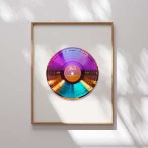 May include: A framed wall art print featuring a circular, iridescent vinyl record graphic with a central label displaying the letters J&B and the text "happily ever after". The record surface includes handwritten text along the outer edge.
