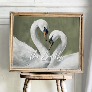 May include: A framed artwork featuring two white swans with curved necks, forming a heart shape. The painting has a green background and includes the text "The Smiths Est. 2026" in a cursive font. The frame is a light brown color.