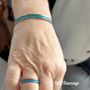 以下が含まれることがあります： 銀色のブレスレットとリングのセットで、どちらもトルコ石がはめ込まれています。リングは指に、ブレスレットは手首に着用されています。ジュエリーは明るく磨かれた仕上がりです。画像には「DFB-Tournage」の文字が含まれています。