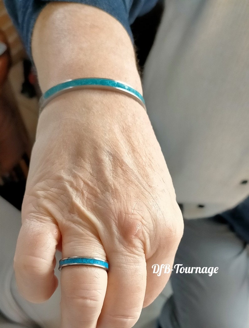 以下が含まれることがあります： 銀色のブレスレットとリングのセットで、どちらもトルコ石がはめ込まれています。リングは指に、ブレスレットは手首に着用されています。ジュエリーは明るく磨かれた仕上がりです。画像には「DFB-Tournage」の文字が含まれています。
