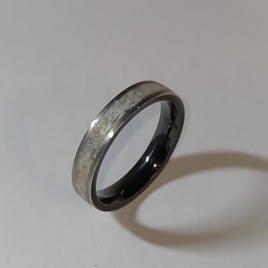 Op de afbeelding: Een zwarte en zilveren ring met een getextureerde, lichtgekleurde inleg. De ring heeft een donker, gepolijst interieur en een zilveren band. De ring is geplaatst tegen een witte achtergrond.