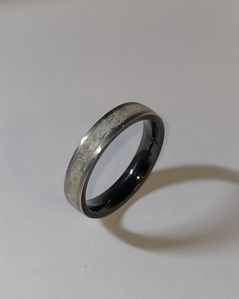 Op de afbeelding: Een zwarte en zilveren ring met een getextureerde, lichtgekleurde inleg. De ring heeft een donker, gepolijst interieur en een zilveren band. De ring is geplaatst tegen een witte achtergrond.