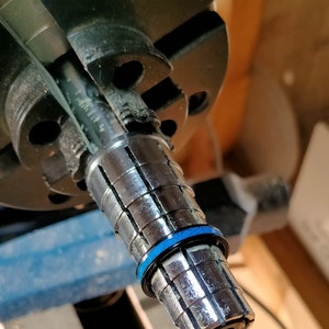 以下が含まれることがあります： セグメント化された円筒形デザインの金属工具のクローズアップ。工具の端近くには青いリングがあり、より大きな黒い機械部品に取り付けられています。背景はぼやけており、ワークショップの環境を示唆しています。