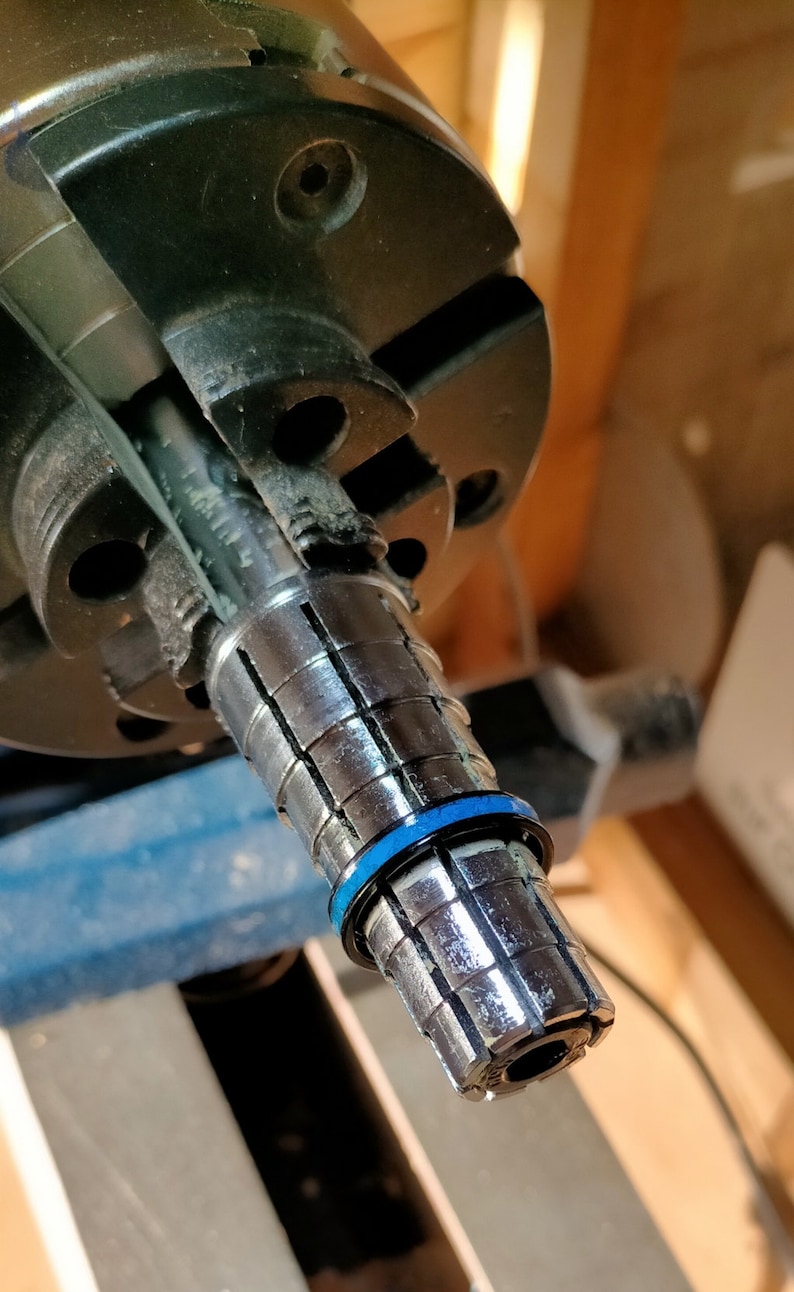 以下が含まれることがあります： セグメント化された円筒形デザインの金属工具のクローズアップ。工具の端近くには青いリングがあり、より大きな黒い機械部品に取り付けられています。背景はぼやけており、ワークショップの環境を示唆しています。