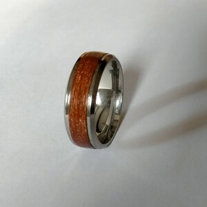 May include: A silver ring with a band of reddish-brown wood inlay. The ring has a polished, metallic finish and a smooth, rounded profile. The wood grain is visible, adding a natural element to the jewelry.