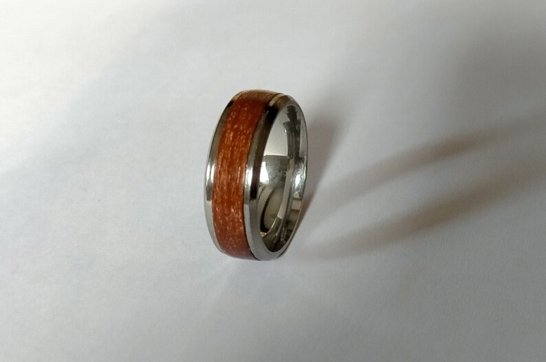 May include: A silver ring with a band of reddish-brown wood inlay. The ring has a polished, metallic finish and a smooth, rounded profile. The wood grain is visible, adding a natural element to the jewelry.