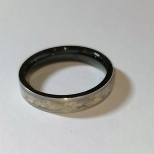 Op de afbeelding: Een zilverkleurige ring met een zwarte binnenband. De ring heeft een getextureerde, lichtgekleurde band in het midden. De ring is waarschijnlijk gemaakt van metaal en is ontworpen om aan een vinger te worden gedragen. De ring ligt op een witte ondergrond.