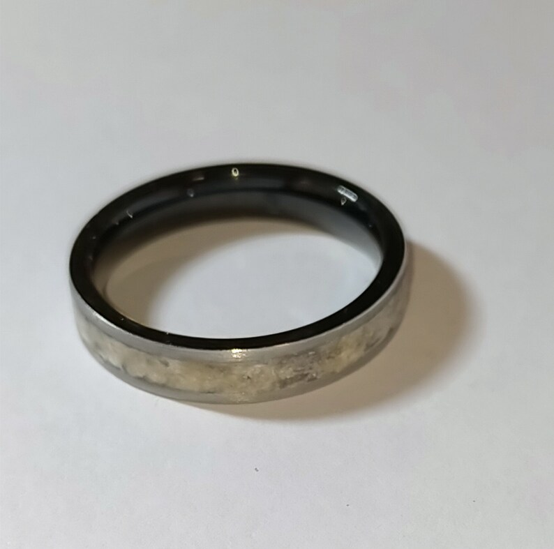 Op de afbeelding: Een zilverkleurige ring met een zwarte binnenband. De ring heeft een getextureerde, lichtgekleurde band in het midden. De ring is waarschijnlijk gemaakt van metaal en is ontworpen om aan een vinger te worden gedragen. De ring ligt op een witte ondergrond.