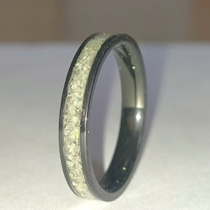 May include: A black ring with a band of light-coloured, textured material. The ring has a sleek, polished appearance, with the light-coloured material set within the black band. The ring is a simple, elegant piece of jewellery.