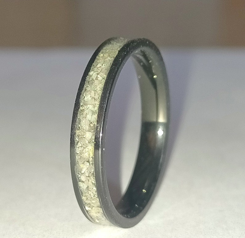 May include: A black ring with a band of light-coloured, textured material. The ring has a sleek, polished appearance, with the light-coloured material set within the black band. The ring is a simple, elegant piece of jewellery.