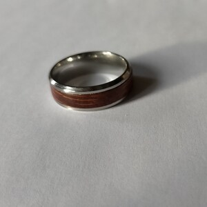 May include: A silver ring with a wide band of reddish-brown wood inlay. The ring has a polished silver finish and a simple, elegant design. The wood grain is visible, adding a natural element to the jewelry.