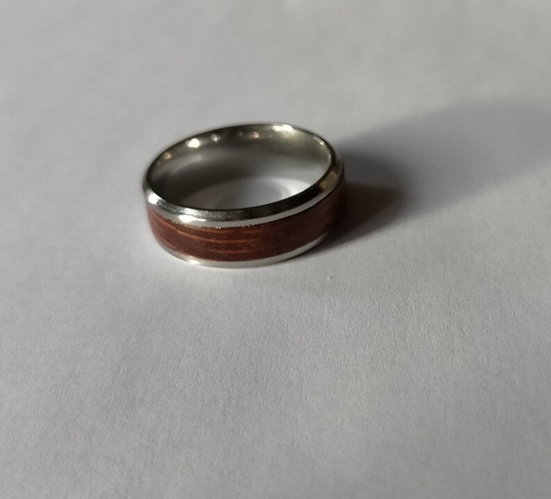 May include: A silver ring with a wide band of reddish-brown wood inlay. The ring has a polished silver finish and a simple, elegant design. The wood grain is visible, adding a natural element to the jewelry.