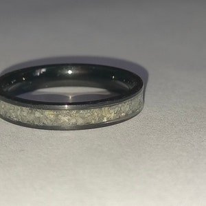 May include: A black ring featuring a band of light-coloured, textured material. The ring is circular and rests on a white surface. The ring's design is simple and modern, suitable for everyday wear.