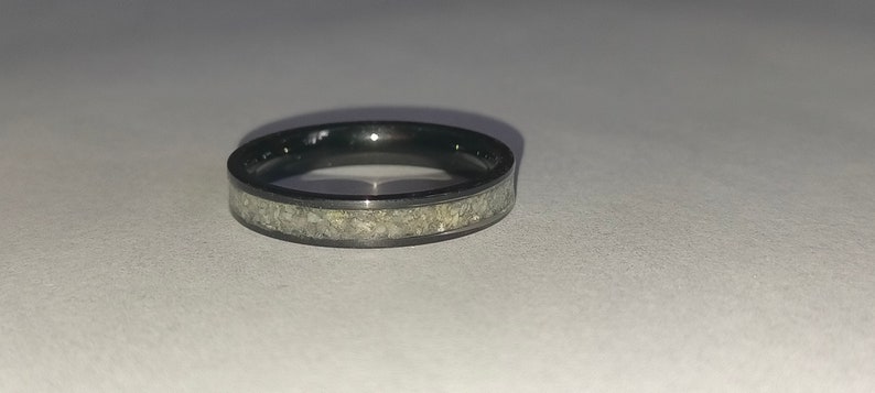 May include: A black ring featuring a band of light-coloured, textured material. The ring is circular and rests on a white surface. The ring's design is simple and modern, suitable for everyday wear.