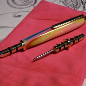 Peut inclure: Un stylo décoratif et un outil assorti avec des accents dorés. Le stylo présente un design coloré avec des teintes bleues, jaunes et rouges. L'outil a une pointe argentée et un manche doré. Les deux articles sont posés sur une surface rose.