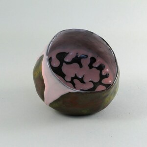 Könnte beinhalten: Handgefertigter Keramikring mit abstraktem Design. Der Ring hat eine dunkelbraune Außenseite, eine rosa Innenseite mit schwarzen Mustern und einen silberfarbenen Rand. Die Form des Rings ist organisch und asymmetrisch, was ihn zu einem besonderen Schmuckstück macht.