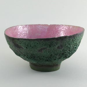 Könnte beinhalten: Handgefertigte Keramikschale mit strukturierter, grüner Außenseite und rosa Innenraum. Die Schale hat einen dunklen Rand und einen kleinen, runden Sockel. Die einzigartige Textur und Farbkombination der Schale machen sie zu einem dekorativen Stück.
