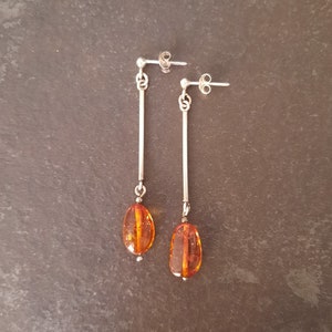 May include: A pair of drop earrings featuring a silver-toned post and a long, thin vertical bar connected to a polished, translucent amber-colored oval bead. The earrings have a secure butterfly back closure and a simple, elongated design.