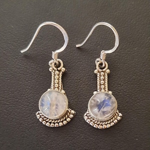May include: A pair of silver-toned earrings with a teardrop design. Each earring features a round, iridescent stone set in a decorative bezel. The earrings have a hook closure.
