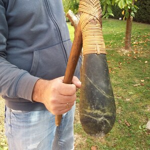 May include: A person holds a dark green stone weapon with a wooden handle wrapped in woven brown material. The weapon is shaped like a club with a rounded end.
