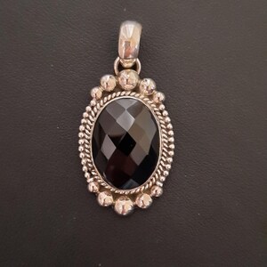 May include: An oval pendant featuring a faceted black gemstone set in a silver frame. The setting includes a twisted rope border surrounded by a series of small, polished silver spheres. A smooth, polished silver bail is attached to the top.