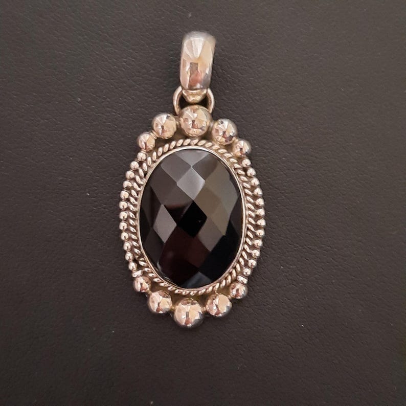 May include: An oval pendant featuring a faceted black gemstone set in a silver frame. The setting includes a twisted rope border surrounded by a series of small, polished silver spheres. A smooth, polished silver bail is attached to the top.
