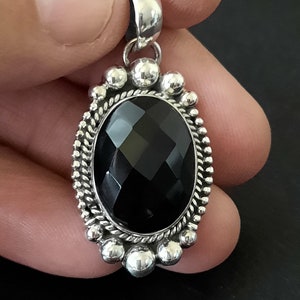 May include: An oval pendant featuring a faceted black gemstone set in a silver frame. The setting includes a twisted rope border surrounded by a series of small, polished silver spheres. A smooth silver bail is attached to the top of the pendant.
