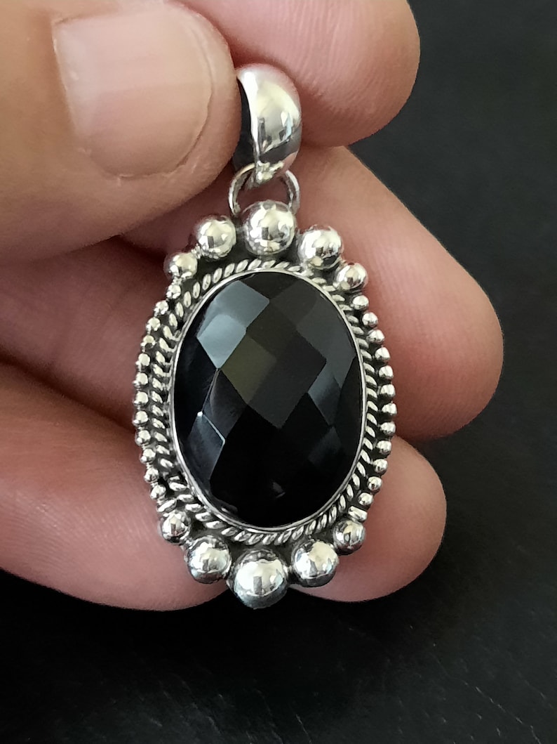 May include: An oval pendant featuring a faceted black gemstone set in a silver frame. The setting includes a twisted rope border surrounded by a series of small, polished silver spheres. A smooth silver bail is attached to the top of the pendant.
