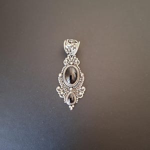 May include: A silver pendant featuring an oval black gemstone at the center and a smaller teardrop-shaped black gemstone below it. The metalwork includes intricate filigree, scroll patterns, and small silver beads surrounding the stones.