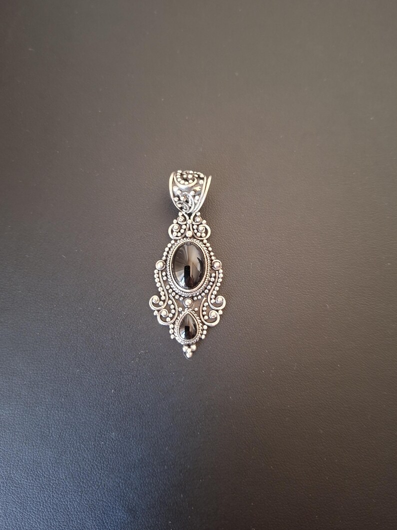 May include: A silver pendant featuring an oval black gemstone at the center and a smaller teardrop-shaped black gemstone below it. The metalwork includes intricate filigree, scroll patterns, and small silver beads surrounding the stones.