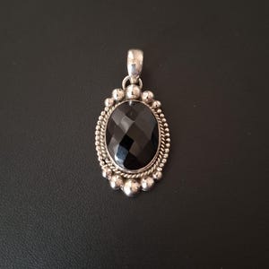 May include: An oval pendant featuring a faceted black gemstone set in a silver frame. The setting is decorated with a twisted rope border and a series of small silver spheres around the edge, topped with a polished silver bail for a chain.