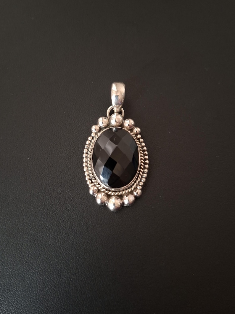 May include: An oval pendant featuring a faceted black gemstone set in a silver frame. The setting is decorated with a twisted rope border and a series of small silver spheres around the edge, topped with a polished silver bail for a chain.