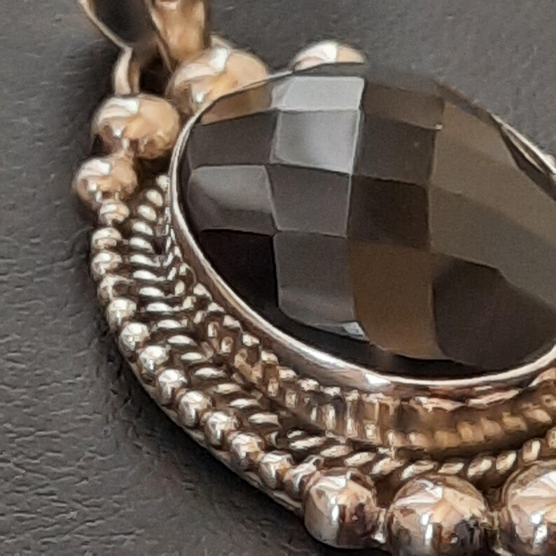 May include: A silver pendant featuring a large, oval, faceted black gemstone set in a metallic frame. The border of the setting is decorated with a textured, beaded, and twisted rope design, with a small loop at the top for attachment to a chain.