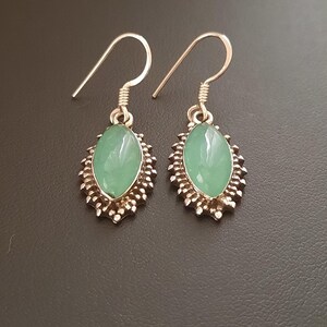 May include: A pair of silver earrings with green oval-shaped stones set in a decorative silver frame. The earrings have a hook closure.