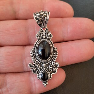Puede incluir: Un colgante de plata con un dise&ntilde;o adornado con dos piedras preciosas negras. El colgante tiene una anilla en la parte superior para sujetarlo a una cadena.