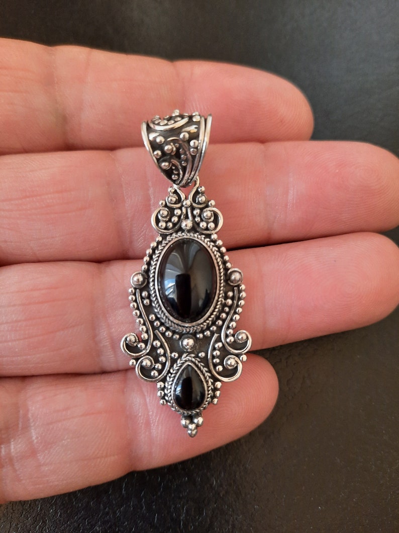 Puede incluir: Un colgante de plata con un dise&ntilde;o adornado con dos piedras preciosas negras. El colgante tiene una anilla en la parte superior para sujetarlo a una cadena.