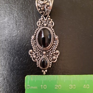 Puede incluir: Un colgante de plata con un dise&ntilde;o adornado presenta dos piedras preciosas negras. El colgante mide aproximadamente 5 cm de largo.