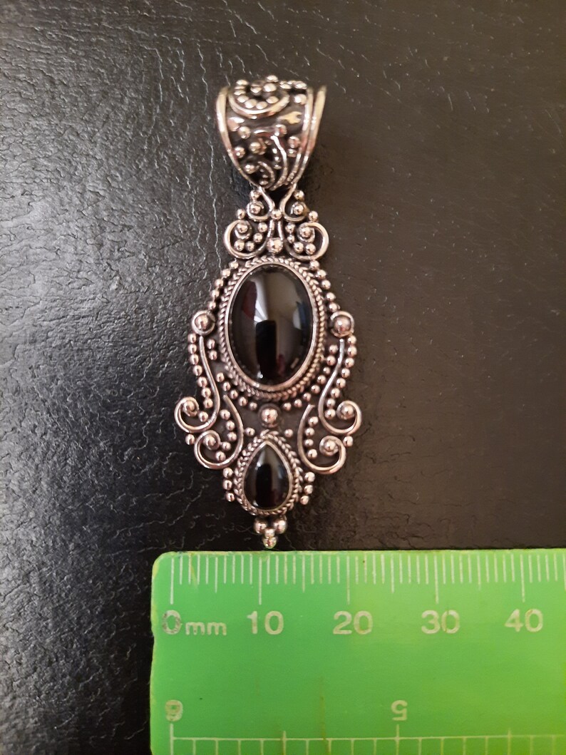 Puede incluir: Un colgante de plata con un dise&ntilde;o adornado presenta dos piedras preciosas negras. El colgante mide aproximadamente 5 cm de largo.