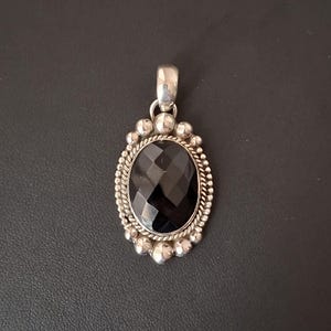May include: An oval pendant featuring a faceted black gemstone set in a silver frame. The setting includes a twisted rope border surrounded by small silver spheres, with a smooth silver bail at the top for attaching to a chain or necklace.
