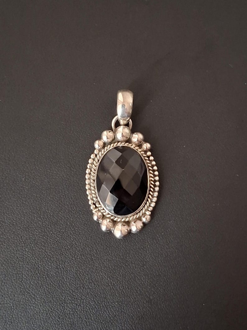 May include: An oval pendant featuring a faceted black gemstone set in a silver frame. The setting includes a twisted rope border surrounded by small silver spheres, with a smooth silver bail at the top for attaching to a chain or necklace.