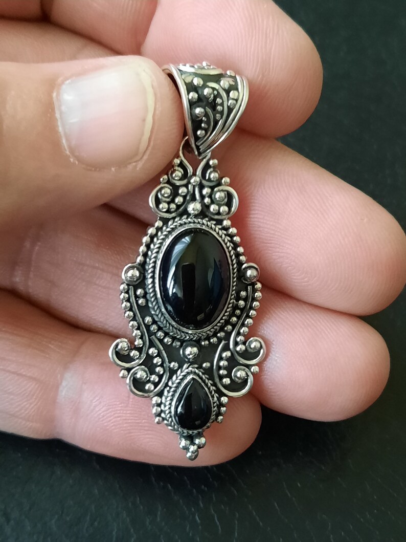 Piedra central ovalada rodeada de filigrana y bolitas de plata de varios tamaños, en la parte inferior piedra en forma de lagrima mas pequeña, con tres bolitas que decoran la farte inferior.