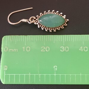 May include: Silver dangle earrings with a green oval gemstone set in a silver filigree design. The earrings measure approximately 2 cm in length.