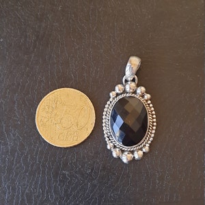 May include: An oval pendant featuring a faceted black gemstone set in a silver frame with a beaded and twisted rope border. A 50 Euro Cent coin is placed next to the pendant for scale, showing the relative size of the jewelry piece.