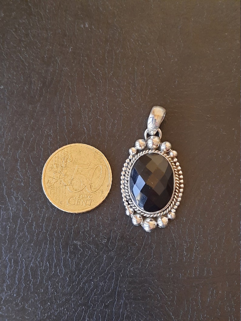 May include: An oval pendant featuring a faceted black gemstone set in a silver frame with a beaded and twisted rope border. A 50 Euro Cent coin is placed next to the pendant for scale, showing the relative size of the jewelry piece.