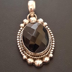 May include: An oval pendant featuring a faceted black gemstone set in a silver frame. The setting includes a twisted rope border surrounded by a series of small, polished silver spheres. A silver bail is attached to the top for hanging.