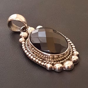 May include: An oval pendant featuring a faceted dark brown gemstone set in a silver frame. The setting is decorated with a twisted rope border and a series of small silver spheres along the outer edge, topped with a polished silver bail.
