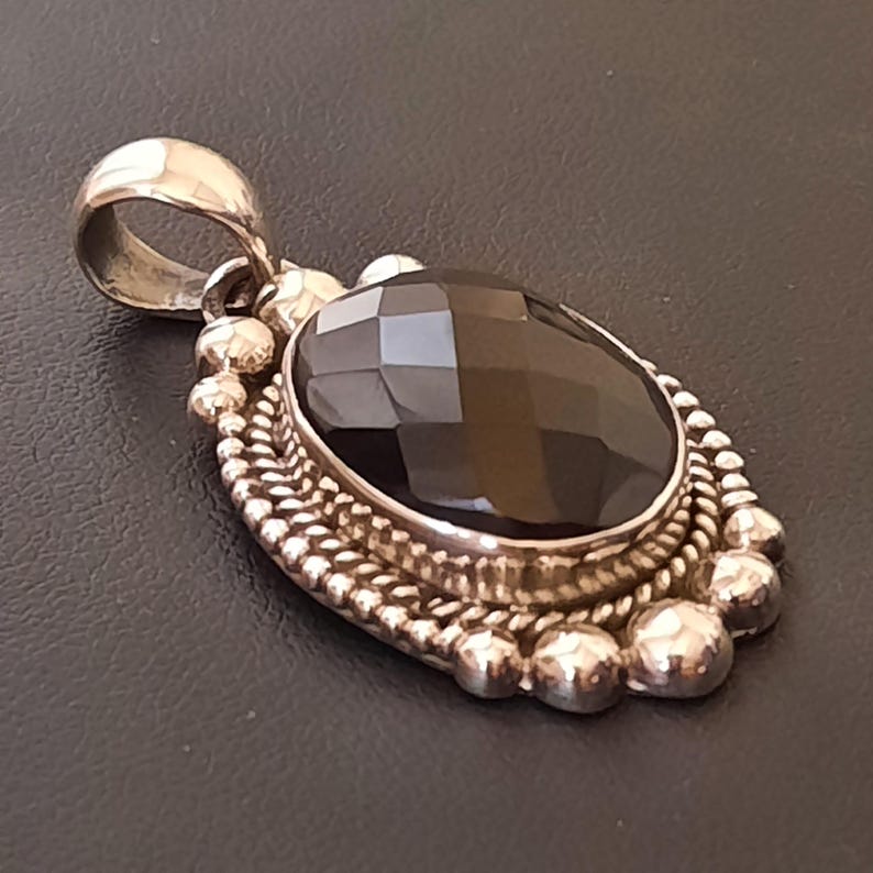May include: An oval pendant featuring a faceted dark brown gemstone set in a silver frame. The setting is decorated with a twisted rope border and a series of small silver spheres along the outer edge, topped with a polished silver bail.