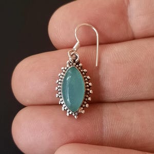 May include: A silver earring with a light blue gemstone set in a decorative silver frame. The earring has a simple hook closure.