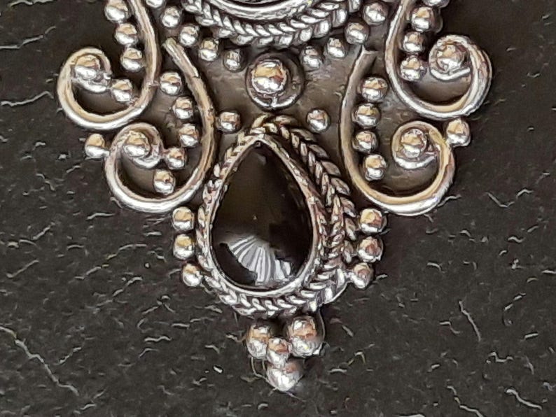 Puede incluir: Un colgante de plata con una piedra preciosa negra en forma de l&aacute;grima. El colgante est&aacute; decorado con intrincados detalles de filigrana.