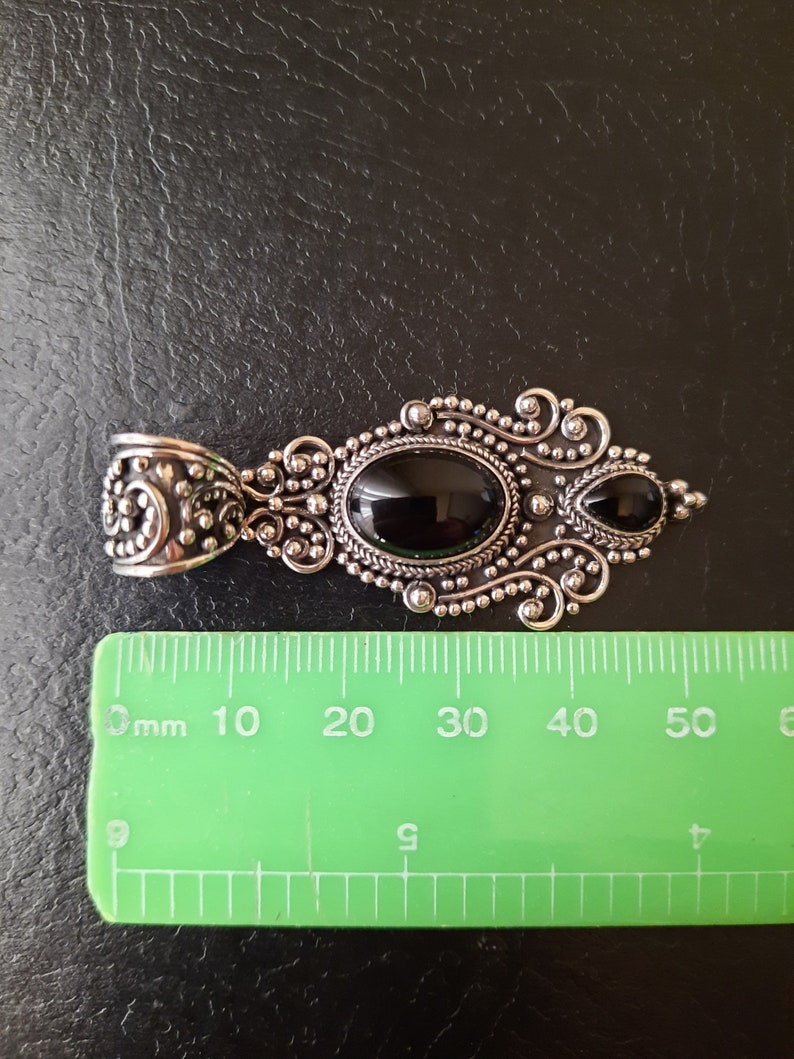 Puede incluir: Un colgante de plata con dos piedras preciosas negras engastadas en un dise&ntilde;o filigranado detallado. El colgante mide aproximadamente 5 cm de largo.